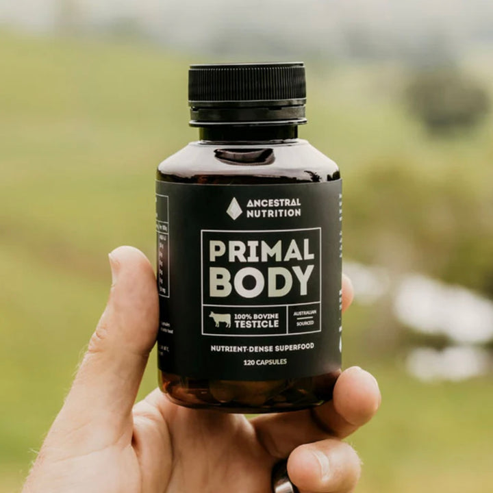 Hand holding a bottle of 'Primal Body' supplement with a blurred natural background