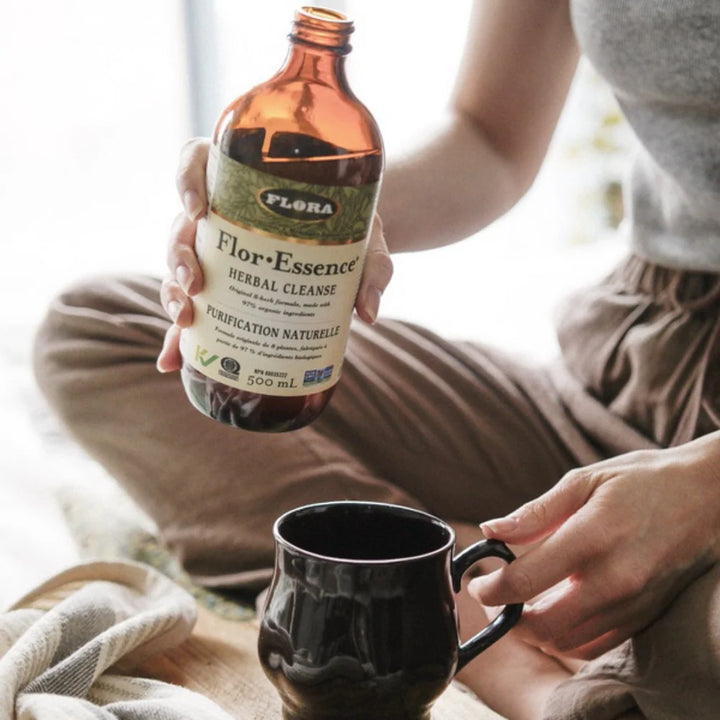 Person holding a bottle of 'Flora Flor Essence Herbal Cleanse' and a black mug.