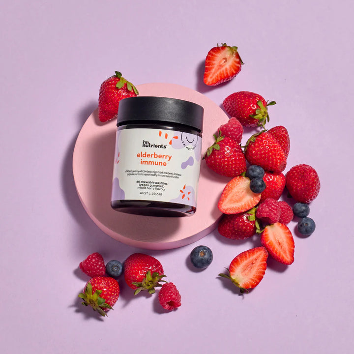 Jar of 'elderberry immune' supplement surrounded by strawberries and blueberries on a pink plate against a light purple background.