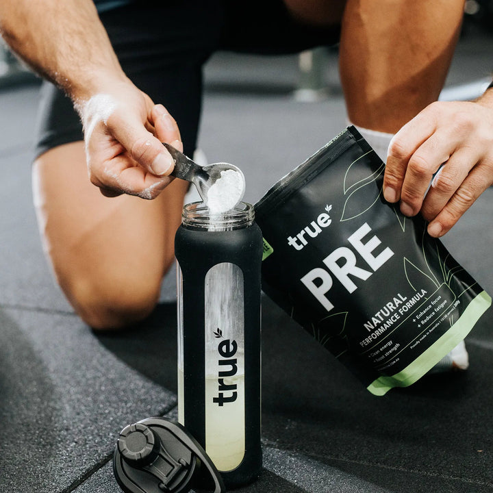 Person preparing a supplement with 'true' brand container and scoop on a gym floor.
