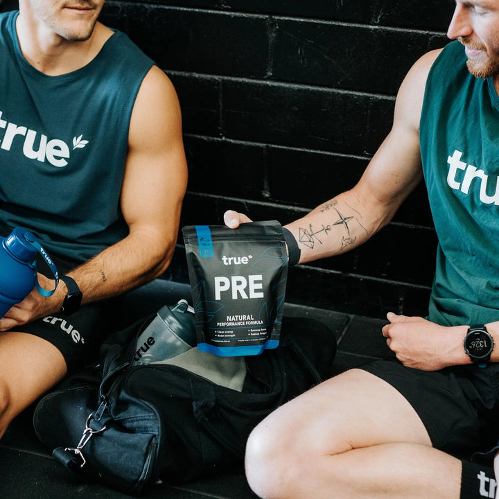 Two men in green 'true' branded sports attire sitting on a black brick wall holding a 'true' pre-workout supplement bag.